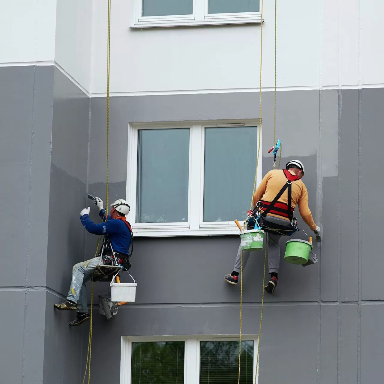 Dois profissionais pintando a fachada de um prédio suspensos por cordas de rapel